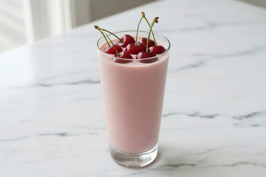 A glass of creamy pink cherry collagen smoothie topped with fresh cherries on a marble counter